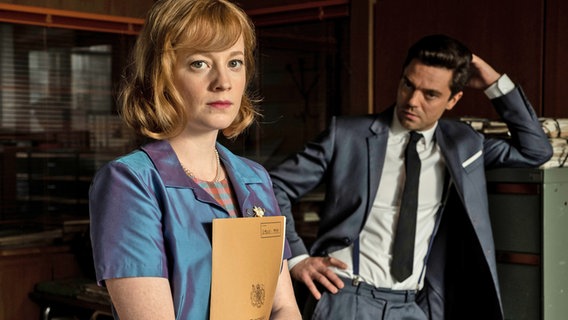Die Spionage-Serie "Spy City" im ZDF - In einem Büro: Elisa Hahn (Leonie Benesch) hält eine Akte in den Händen und Fielding Scott (Dominic Cooper) stützt sich auf einem Aktenschrank ab. © ZDF und 2020 Odeon Fiction Foto: Dušan Martinček.