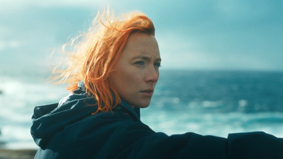 eine junge Frau mit roten wehenden Haaren und blauen Augen schaut skeptisch, hinter ihr tost das Meer - Szene aus "the Outrun" mit Saoirse Ronan © The Outrun Ltd. 