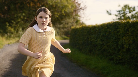 Ein Mädchen in gelbem Kleid und wehenden Haaren läuft einen Weg hinunter © Neue Visionen Filmverleih 