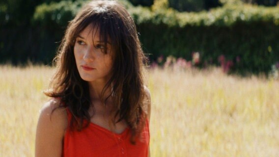 Eine Frau in einem roten Sommerkleid schaut verträumt, während sie über ein Feld geht. © Les Films Pelléas - Année Zéro 