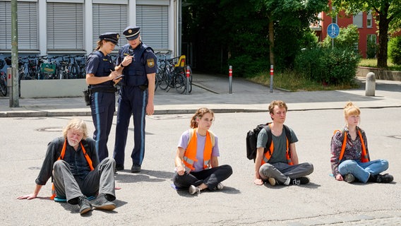 Ein Polizist spricht in sein Funkgerät, während eine andere Polizistin etwas auf einem Block notiert. Beide stehen hinter Protestierenden, die nebeneinander auf der Straße sitzen und Warnwesten tragen. © Honorarfrei - nur für diese Sendung inkl. SocialMedia bei Nennung ZDF und Bernd Schuller 