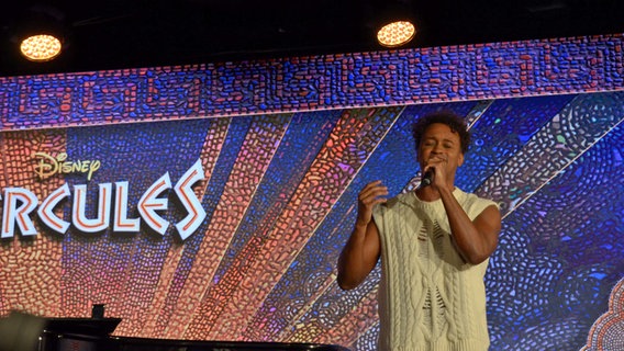 Der brasilianische Sänger Bénet Monteiro singt mit einem Mikrofon vor der Kulisse mit der Aufschrift "Hercules" © NDR.de Foto: Patricia Batlle
