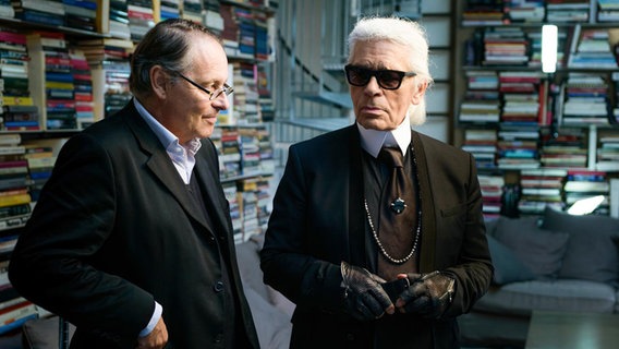 Karl Lagerfeld und ein zweiter Mann stehen in einem Raum voller Regalen mit Musterbüchern und Lagerfeld mustert den Mann skeptisch durch die Sonnenbrille. © 3 Sat / ZDF 