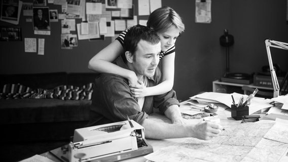 Ein Frau umarmt von hinten einen Mann, der am Schreibtisch sitzt und sich eine Landkarte ansieht. © Zeitsprung Pictures / Wild Bunch Germany Foto: Peter Hartwig
