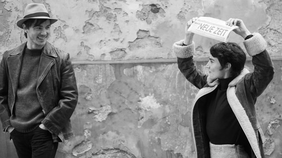 Ein Mann mit Hut steht vor einer maroden Mauer und lächelt eine Frau an, die eine Zeitung in die Luft hält. © Zeitsprung Pictures / Wild Bunch Germany Foto: Peter Hartwig
