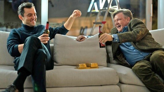 Der junge Anwalt Sven Lebert (Nils Strunk) und sein Chef Dr. Bernd Hausner (Justus von Dohnányi) sitzen auf einem Sofa und prosten sich erfreut mit einer Flasche Bier zu. © X Filme Creative Pool GmbH / True Content Entertainment ApS / EPO-Film / ZDF / DR / Petro Domenigg 