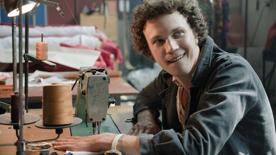 Renars (Karlis Arnolds Avots) sitzt grinsend an einer Industrienähmaschine und will Jeans in der Sowjetunion produzieren. © Arte, TASSE FILM 
