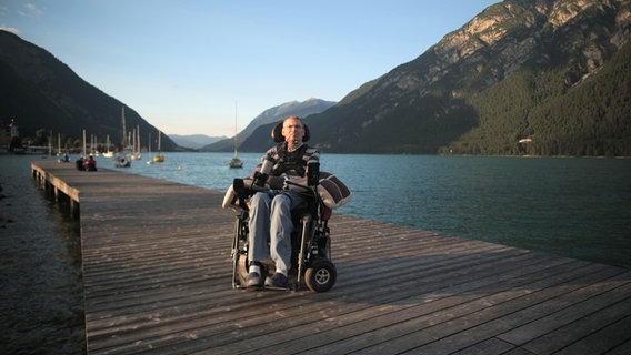 Ein Mann im Rollstuhl vor einem See und einem Bergpanorama © NDR/Torsten Lapp 