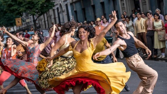 Mehrere junge Leute in beiger und dunkler Kleidung laufen auf einer Straße vor einer Fabrikhalle entlang - Szene aus dem Film "West Side Story" von Steven Spielberg © 2020 Twentieth Century Fox Film Corporation. Foto: Niko Tavernise