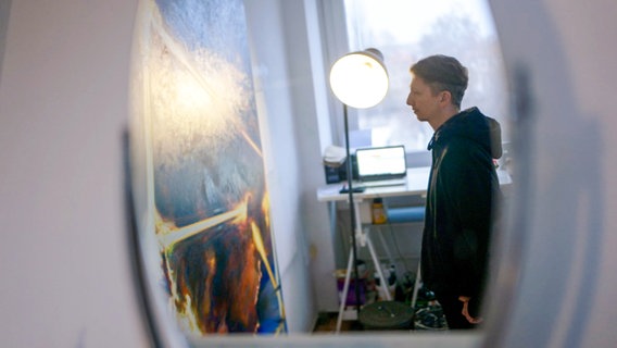 Ein Mann steht vor einem Gemälde und wird durch eine Lampe angestrahlt. © NDR 