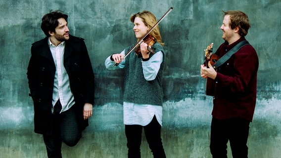 Das Trio Stundom posiert vor einer Wand, eine Musikerin hält eine Geige in der Hand. © Annette Kjær Foto: Annette Kjær