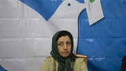 Eine Frau mit Kopftuch sitzt vor einem Plakat mir einer Friedenstaube. © picture alliance / NurPhoto | Morteza Nikoubazl 