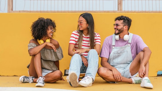 Drei jungen Menschen sitzen vor einer gelben Wand und lachen sich an. © IMAGO / Addictive Stock 