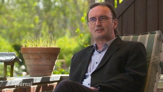 Gerhard Henschel sitzt während eines Interviews auf einem Gartenstuhl auf einer Terrasse.  Foto: Screenshot