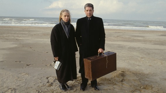 Leonard Cohen und Dominique Isserman posieren an einem Strand, er hat einen Koffer und sie eine Filmklappe in der Hand. © © 2022 PROKINO/ Eric Préau/Sygma via Getty Images 