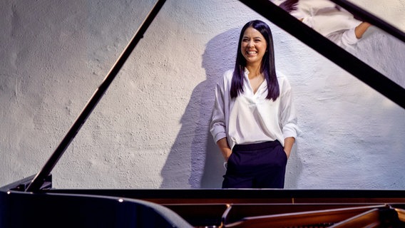 Die Pianistin Hanni Liang steht mit ihren Händen in den Hosentaschen hinter einem Flügel und lächelt. © Felix Broede Foto: Felix Broede