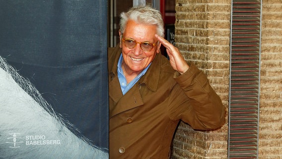 Der Schauspieler Henry Hübchen lächelt und salutiert, indem er seine linke Hand an die Schläfe hält. © picture alliance/dpa | Gerald Matzka 