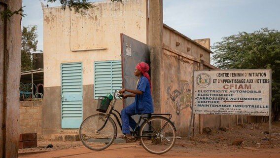 Eine Schwarze Frau mit Blaumann und rotem Turban sitzt gerade auf einem Fahrrad und lächelt. Sie fährt durch ein Tor. © NDR Foto: Sascha Montag