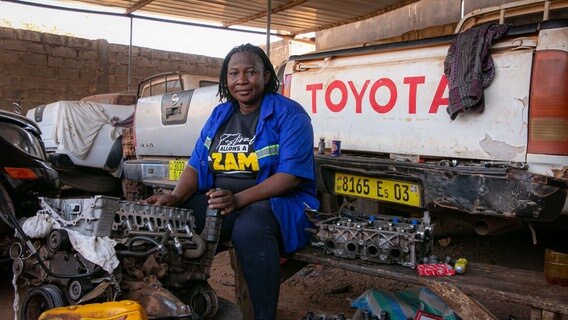 Eine korpulente Frau mit schwarzen Rastalocken arbeitet an einem elektronischen Werkstück. sie sitzt in einem Werkstatt umgeben von Metall, Autoreifen und Autos. © NDR Foto: Sascha Montag