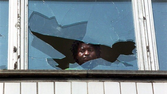 Ein Afrikaner blickt am Tag nach den ausländerfeindlichen Übergriffen in Hoyerswerda (Sachsen) im September 1991 verängstigt durch die eingeworfene Scheibe eines Wohnheims. © picture alliance / dpa Foto: Rainer Weisflog