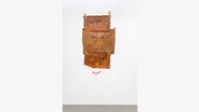 An einer Wand hängen drei braune Tücher, auf denen Augen abgebildet sind. Darunter ist ein roter Mund gemalt. © Harry Thring 