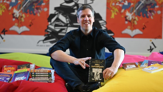 Jeff Kinney sitzt auf dem Boden und hält sein neues Buch "Gregs Tagebuch 17: Voll aufgedreht!" in der Hand. © picture alliance/dpa Foto: Lars Klemmer