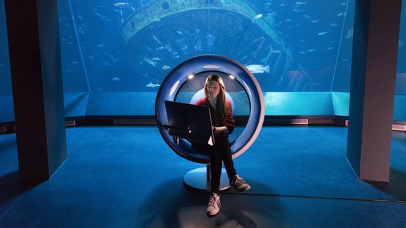 Sound chair with a woman in front of a large aquarium © Anke Neumeister / Deutsches Meeresmuseum Photo: Anke Neumeister