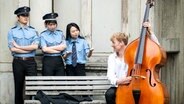 Drei als Polizisten verkleidete Menschen stehen streng blickend hinter einer Parkbank, auf der  Dr. Nepomuk Riva mit einem Kontrabass sitzt und zu ihnen aufschaut. © Isabel Winarsch für VolkswagenStiftung 