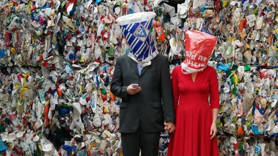 Zwei Menschen mit Plastiktüten über dem Kopf stehen vor einem Plastikmüllberg © Swaantje Güntzel/Scheibe & Güntzel Foto: Scheibe & Güntzel