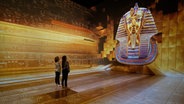 Zwei Menschen stehen in einem gold ausgeleuchtetem Raum und blicken auf eine riesige ägyptische Maske. © MAD 