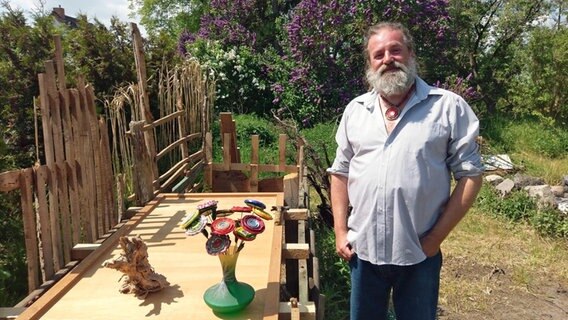 Markus Metz steht in seinem Garten in Altenhagen, neben ihm stehen zwei Objekte seiner Kunst aus Kronkorken auf einem Tisch. © NDR Foto: Lenore Lötsch