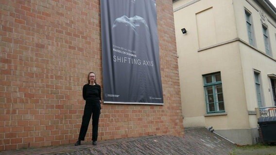 Eine Frau in schwarzer Kleidung steht vor einer Backsteinwand mit einem großen dunklen Bildplakat. © NDR Foto: Helgard Füchsel