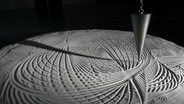 Eine große graue Pendelspitze zeichnet ein ellipsenartiges Muster in eine große Sandfläche. © NDR Foto: Helgard Füchsel