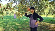Die Künstlerin Natalie Deseke hält ein orangefarbenes Stoffbeutelchen, dass mit einem Seil an einem Baum befestigt ist. © NDR Foto: Agnes Bührig