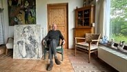 Der Maler Oskar Manigk sitzt einem Wohnzimmer, neben ihm lehnen Bilder. © NDR Foto: Juliane Voigt