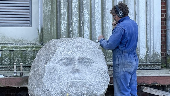 Ein Künstler arbeitet an einer Skulptur aus Stein, die wie ein Gesicht aussieht. © NDR Foto: Frank Hajasch