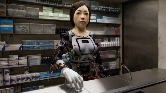 Ein Roboter mit weiblichem Körper steht vor einem Regal voller Arzneimittel. © Dries Verhoeven Foto: Willem Popelier