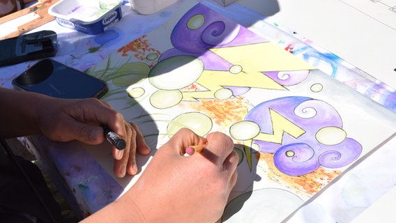 Eine Hand, die im Sonnenlicht gelbe und violette grafische Formen zeichnet - im Open-Air-Atelier zum Gedenken an Wolfgang Herrndorf. © Kunstkreis Norderstedt 