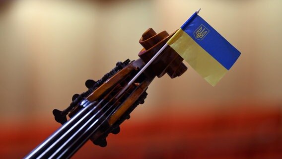 Eine Fahne der Ukraine steckt vor Beginn eines Konzerts an einem Saiten-Instrument des Kyiv Symphony Orchestras. © picture alliance/dpa | Robert Michael 