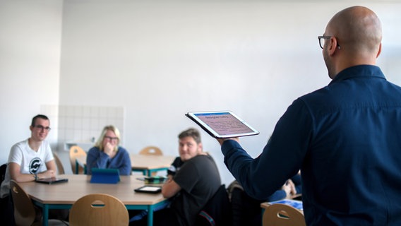 Ein Lehrer steht vor einer Klasse und hält ein iPad in die Luft. © picture alliance/dpa | Britta Pedersen 
