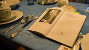 Ein aufgeschlagenes Buch liegt auch einem festlich gedeckten Tisch. © NDR Foto: Tim Piotraschke