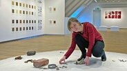 Lena Kaapke kniet inmitten ihrer Ausstellung "Feldforschungen": An den Wänden hinter ihr sind Arbeiten aufgehängt, vor ihr liegen verschiedene Objekte. © NDR Foto: Frank Hajasch