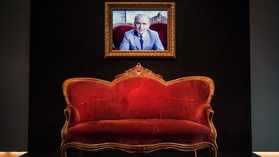 Das rote Sofa, auf dem Vicco von Bülow alias Loriot oft im Fernsehen zu sehen war, steht  im Haus der Geschichte in Stuttgart. Über dem Sofa hängt ein vergoldetet Rahmen, auf dem sein Bild zu sehen ist. © picture alliance / dpa | Wolfram Kastl 