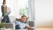 Ein Mann liegt auf dem Sofa und schaut fern, während eine Frau im Hintergrund einen Wäschekorb trägt © Bildagentur-online/Tetra Images 