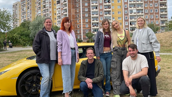 Darsteller und Crew der Produktion "Mels Block" posieren vor einem gelben Sportwagen und einer Hochhaus-Kulisse für ein Gruppenbild. © NDR Foto: Jürn-Jakob Gericke