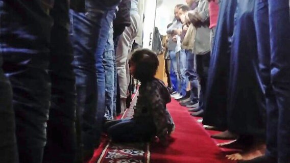 Ein Junge kniet in einer Moschee zwischen stehenden Männdern © NDR 