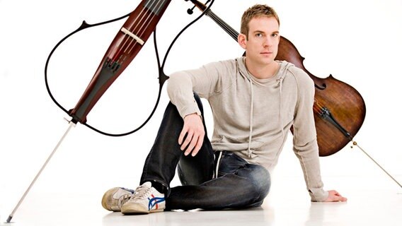 Der Cellist Johannes Moser sitzt vor zwei Instrumenten, einem akkustischem und einem elektrischem Cello.  