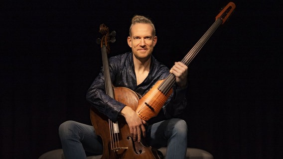 Der Cellist Johannes Moser sitzt vor zwei Instrumenten, einem akkustischem und einem elektrischem Cello. Dabei trägt er eine schwarze Lederjacke und hat blonde, kurze Haare. Er sitzt vor einem schwarzen Hintergrund. © Johannes Moser 
