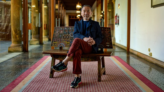 Der Autor Haruki Murakami sitzt mit verschränkten Beinen auf einer Holzbank und blickt in die Kamera. © picture alliance / ASSOCIATED PRESS | Alvaro Barrientos 