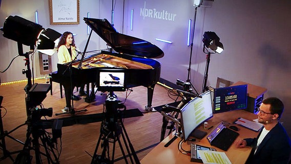 Man sieht ein Studio von NDR Kultur, an einem schwarzen Flügel sitzt eine junge Frau mit braunen, langen und gelockten Haaren, ihr gegenüber sitzt ein Mann an einem Schreibtisch, auf dem zwei Bildschirme stehen. © NDR Screenshots Foto: NDR Screenshots
